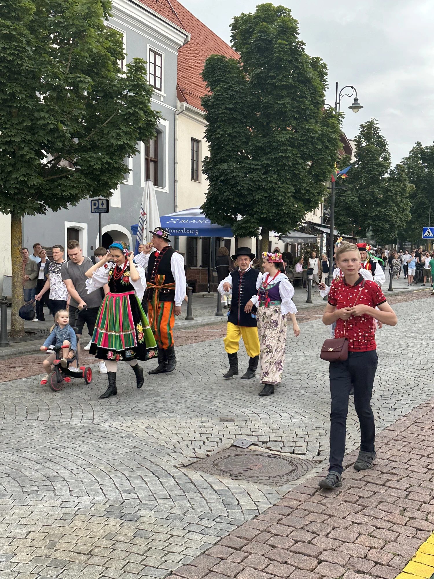 People wearing colorful traditional Baltic or Polish folk costumes walk down a cobblestone street in Vilnius Old Town, with historic buildings and green trees in the background.