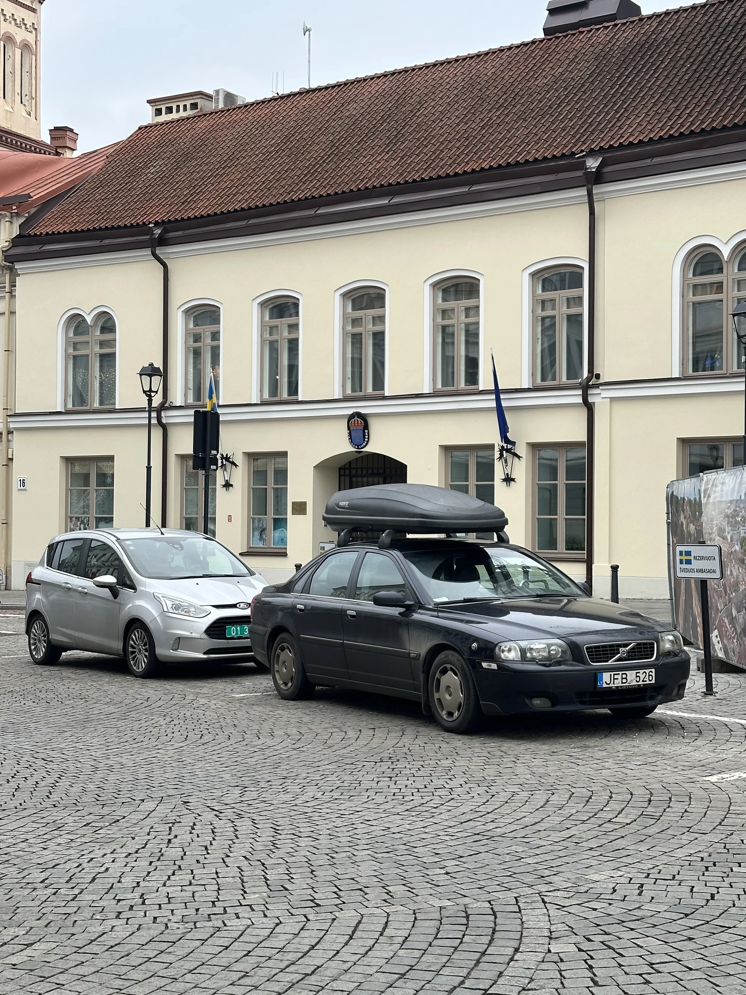 A black Volvo with a roof box and a silver Ford parked on a cobblestone street in front of the pastel-colored Embassy of Sweden building in Vilnius.