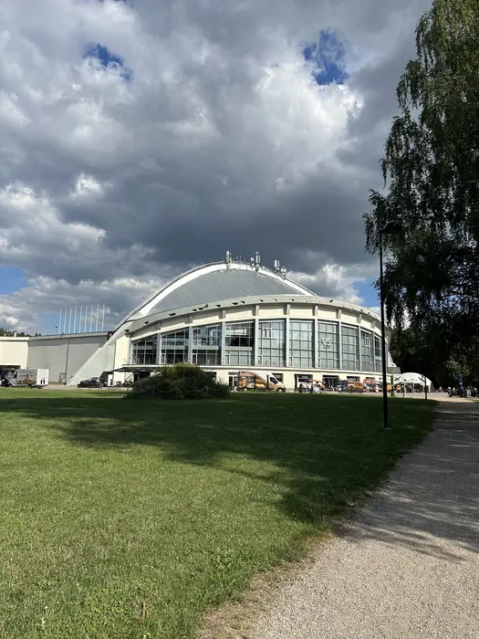 A large dome-shaped building with a glass facade stands next to a wide green lawn and a dirt path under a dramatic cloudy sky in Vingis Park, Vilnius.