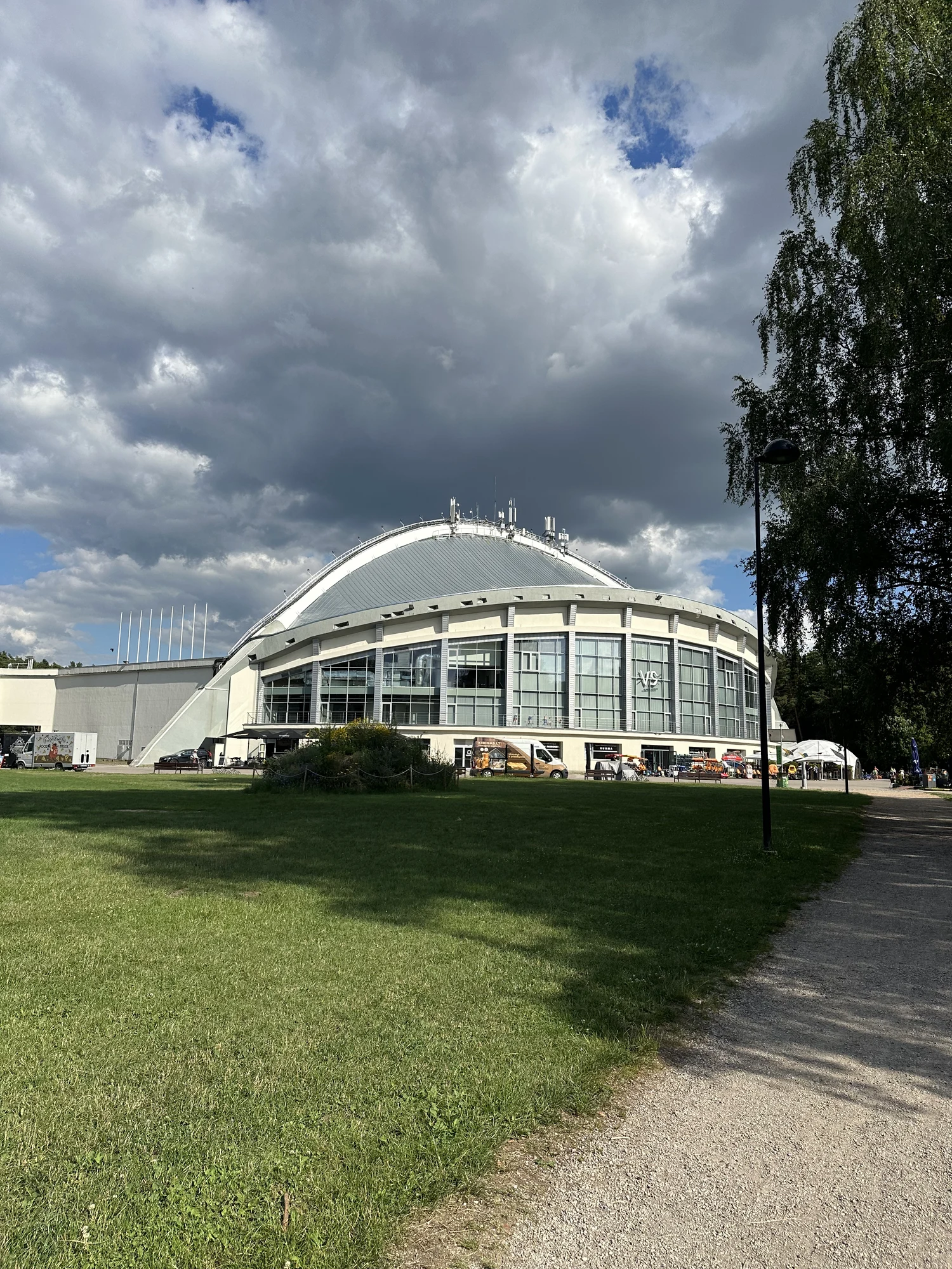 A large dome-shaped building with a glass facade stands next to a wide green lawn and a dirt path under a dramatic cloudy sky in Vingis Park, Vilnius.