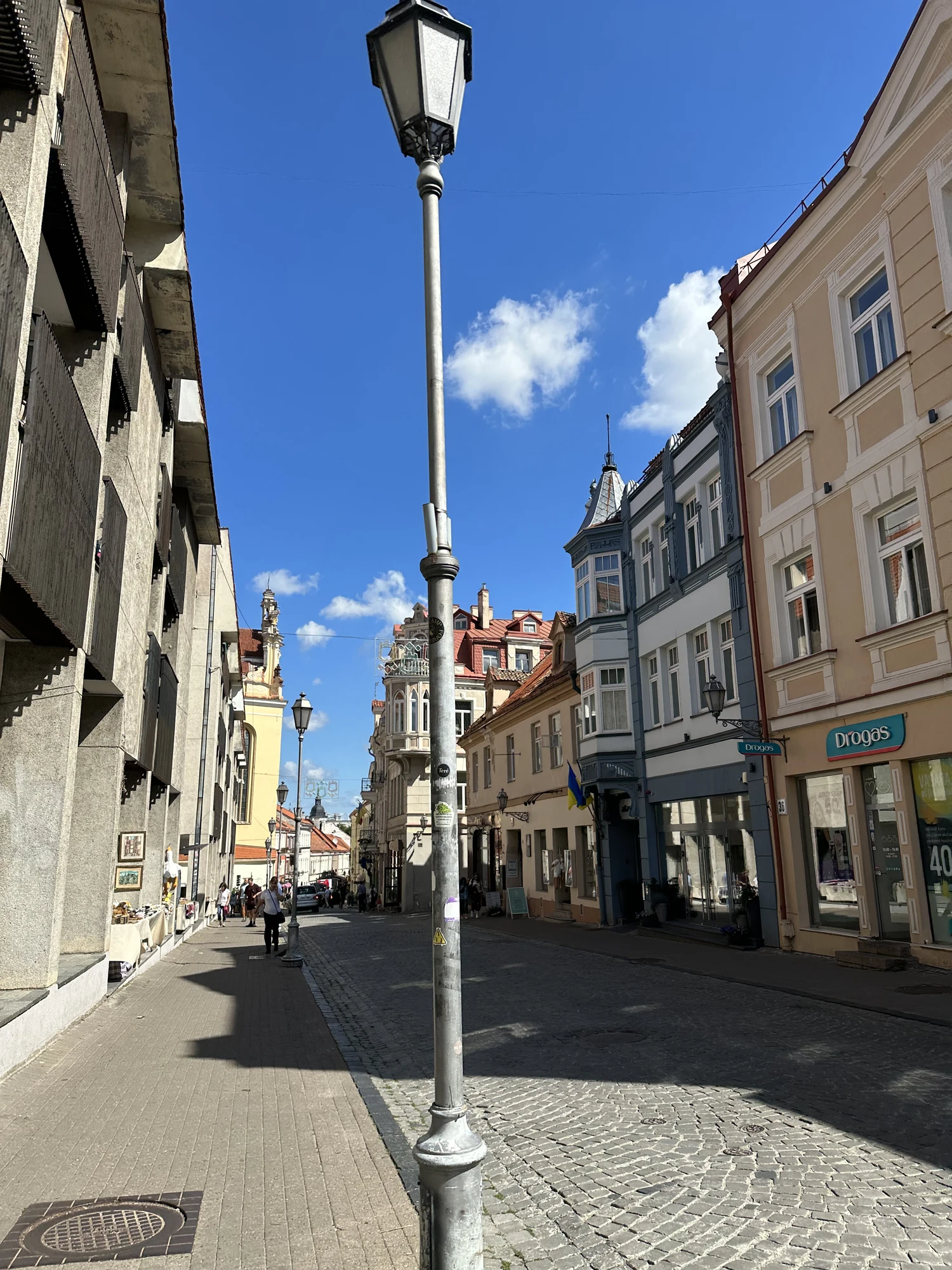 A cobblestone street in Vilnius Old Town lined with historic European buildings, featuring a tall metal lamppost in the foreground under a bright blue sky.