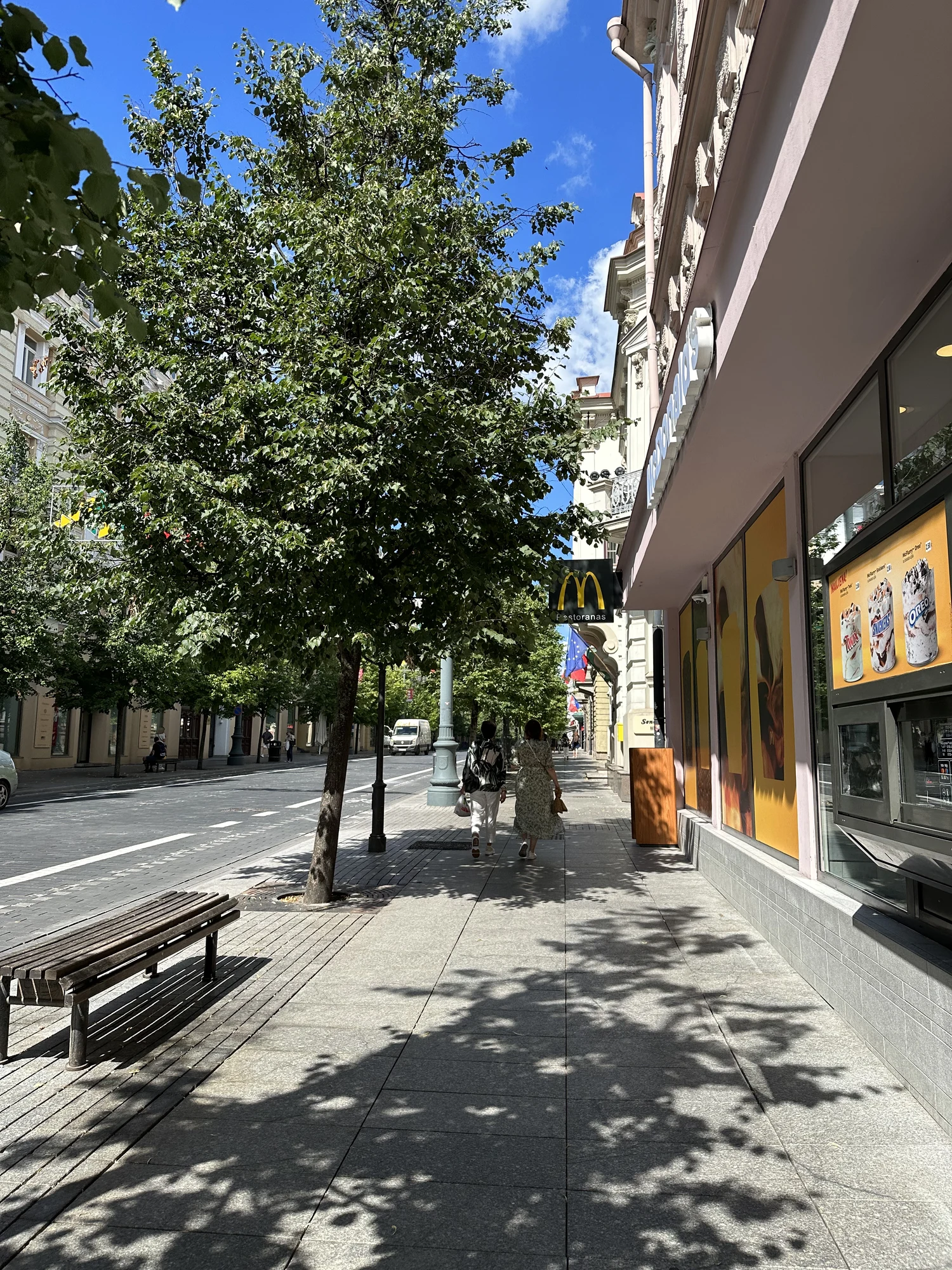 A sunny sidewalk on Gediminas Avenue in Vilnius, with tree shadows cast on the pavement, people walking, and a McDonald's restaurant on the right.