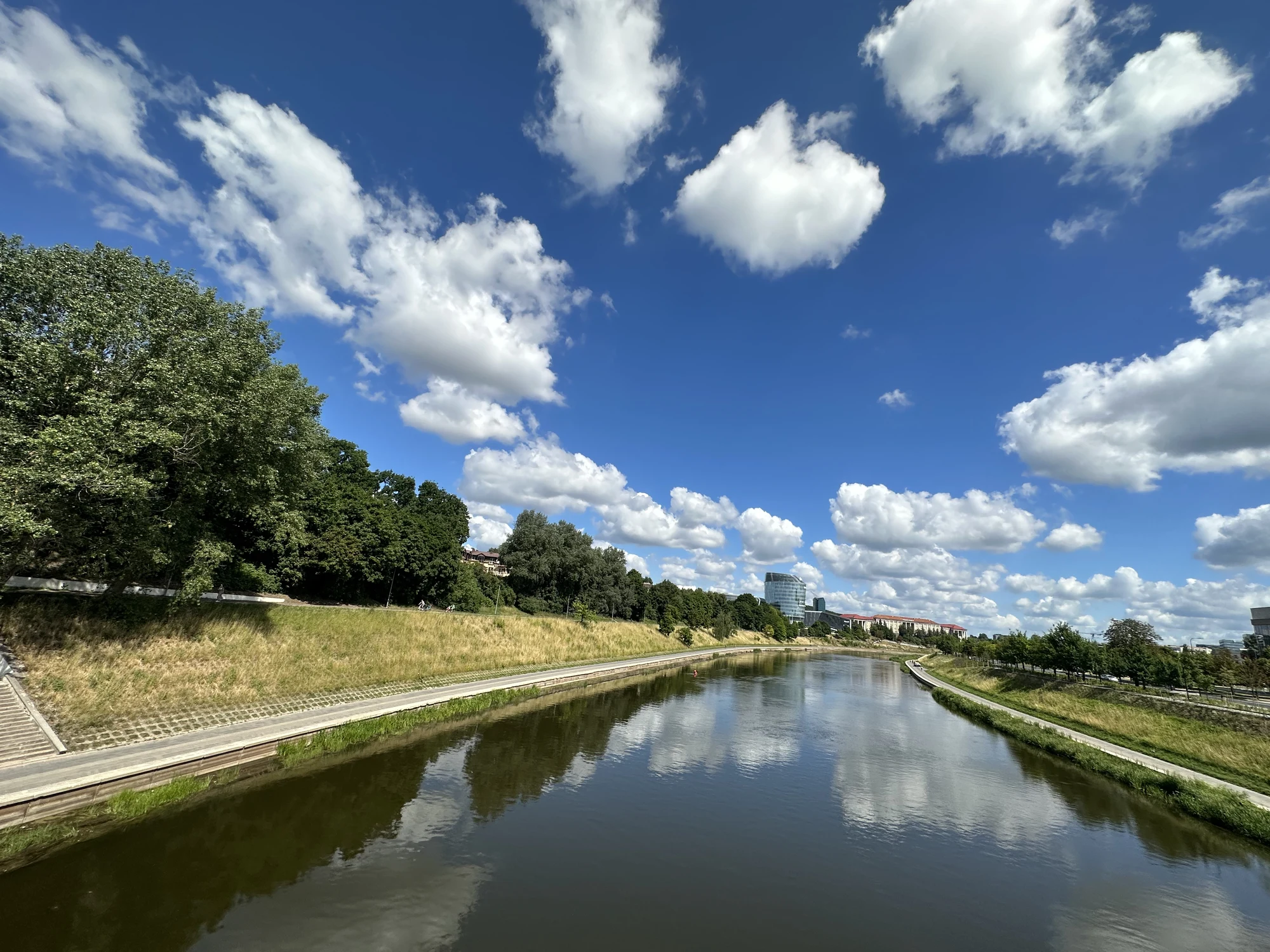 A wide-angle view of the Neris River in Vilnius under a vibrant blue sky with white clouds, featuring grassy banks, trees, and walking paths on both sides.