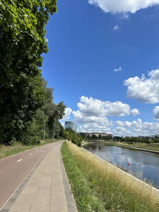 A paved pedestrian and bicycle path runs alongside the Neris River in Vilnius under a bright blue sky with fluffy white clouds.