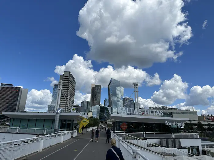 A view from the Baltas Tiltas bridge in Vilnius, showing pedestrians walking towards modern high-rise buildings under a blue sky with large white clouds.
