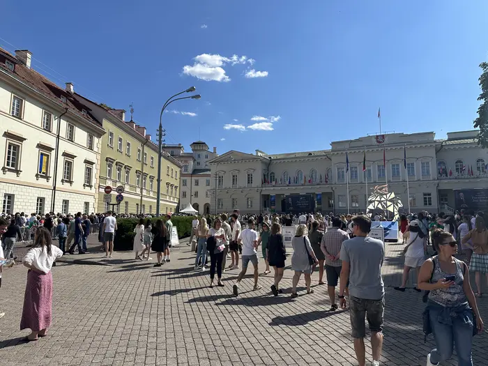 A wide shot of Daukanto Square in Vilnius, showing the Presidential Palace in the background under a blue sky, with many people walking across the paved square in the foreground.