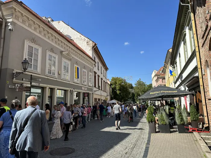 A sunny day on Pilies Street in Vilnius, showing pedestrians walking along the cobblestone road lined with historic buildings, outdoor cafes, and hanging flags.