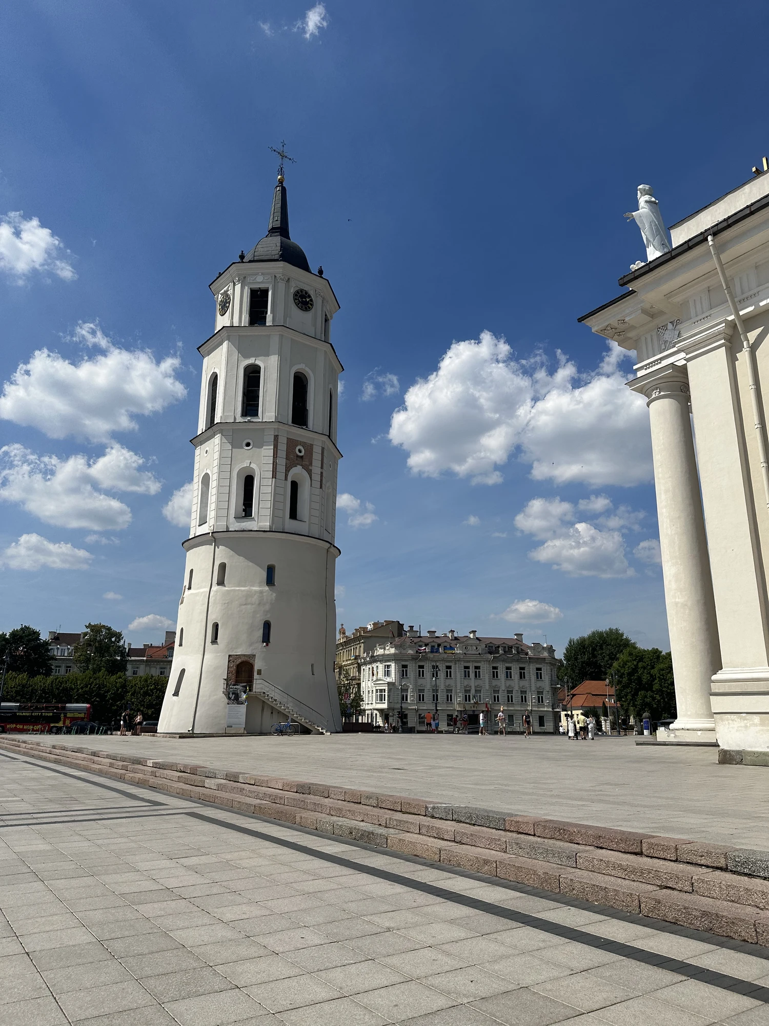 A tall white historic bell tower next to the Vilnius Cathedral on a paved public square under a blue sky with white clouds.
