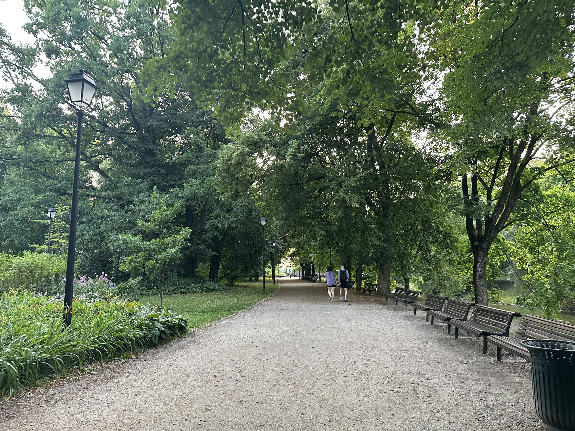 A wide gravel walking path in Bernardine Garden, Vilnius, lined with wooden benches on the right, street lamps, and lush green trees overhead, with two people walking in the distance.