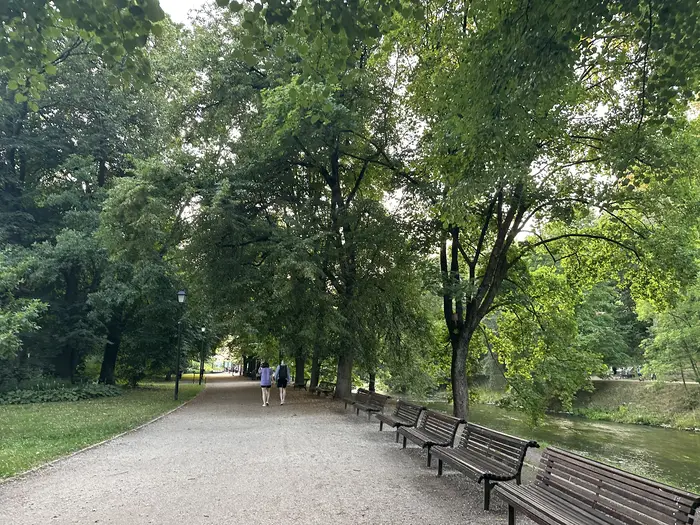 A gravel walking path in a green park lined with wooden benches on the right next to a flowing river, with two people walking in the distance under a canopy of green trees.