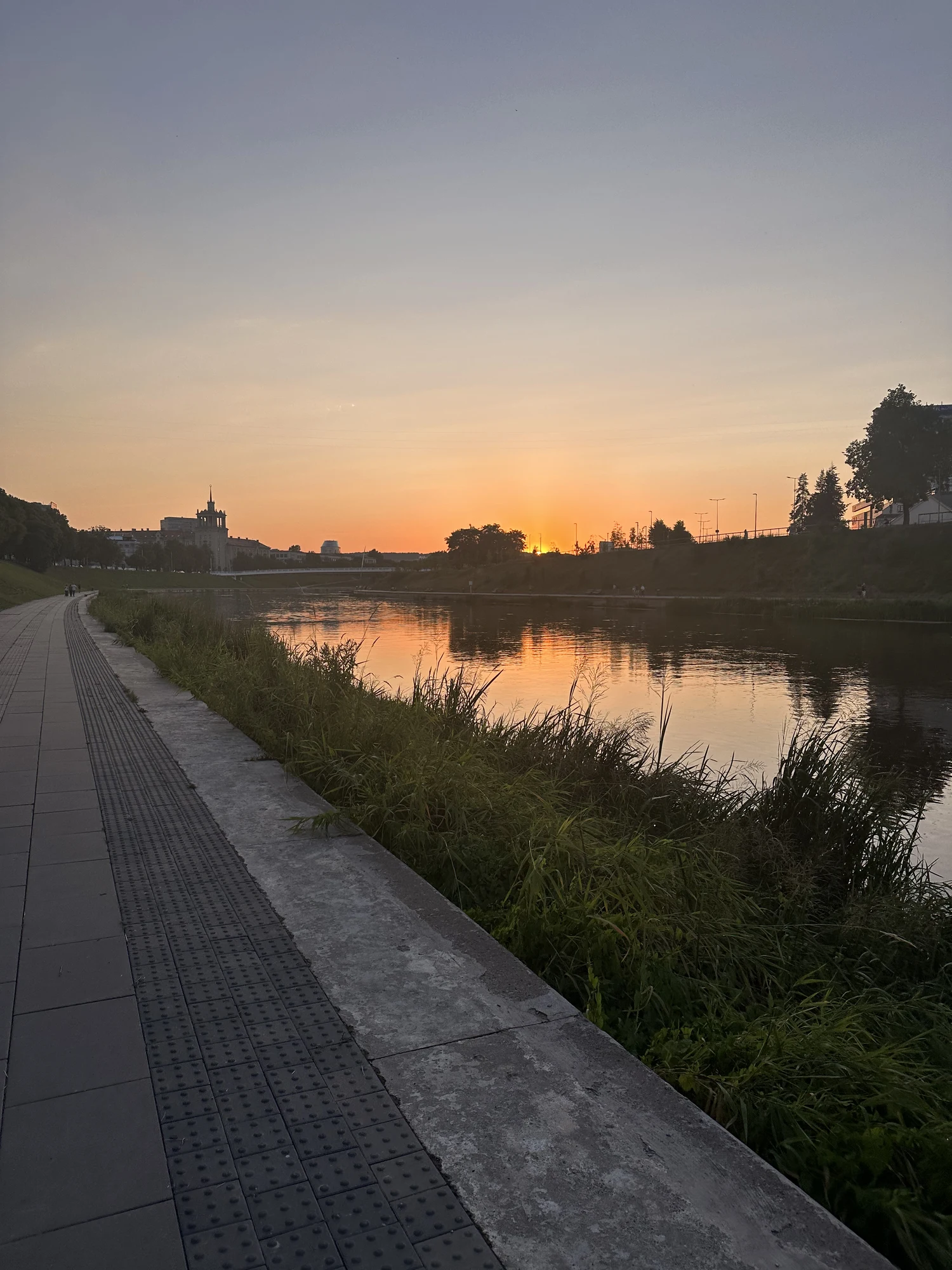 A paved pedestrian path runs along the grassy banks of the Neris River in Vilnius during a vibrant sunset.