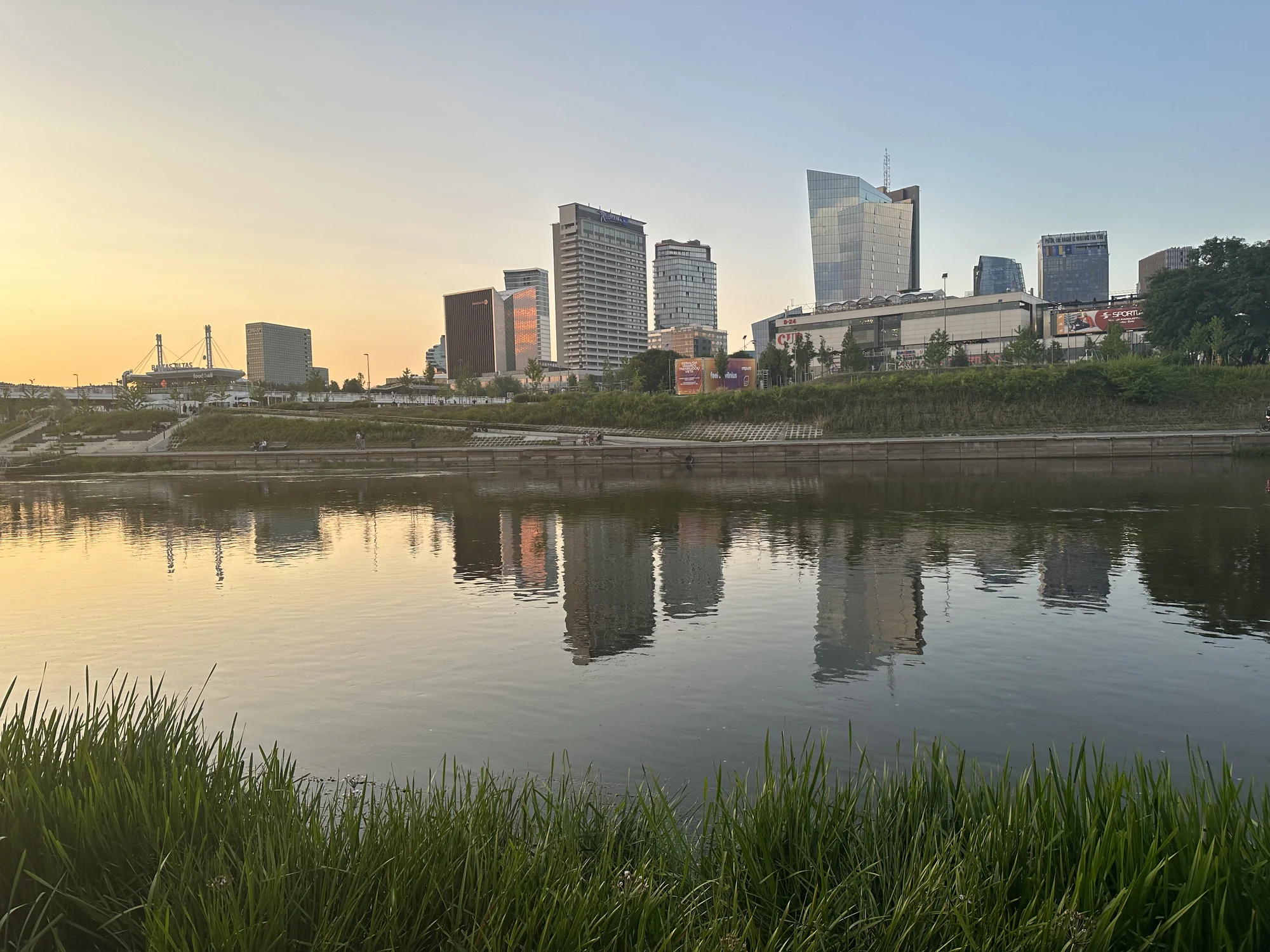 A view across the Neris River in Vilnius showing the modern skyline of high-rise buildings reflected in the water, framed by green grass in the foreground at sunset.
