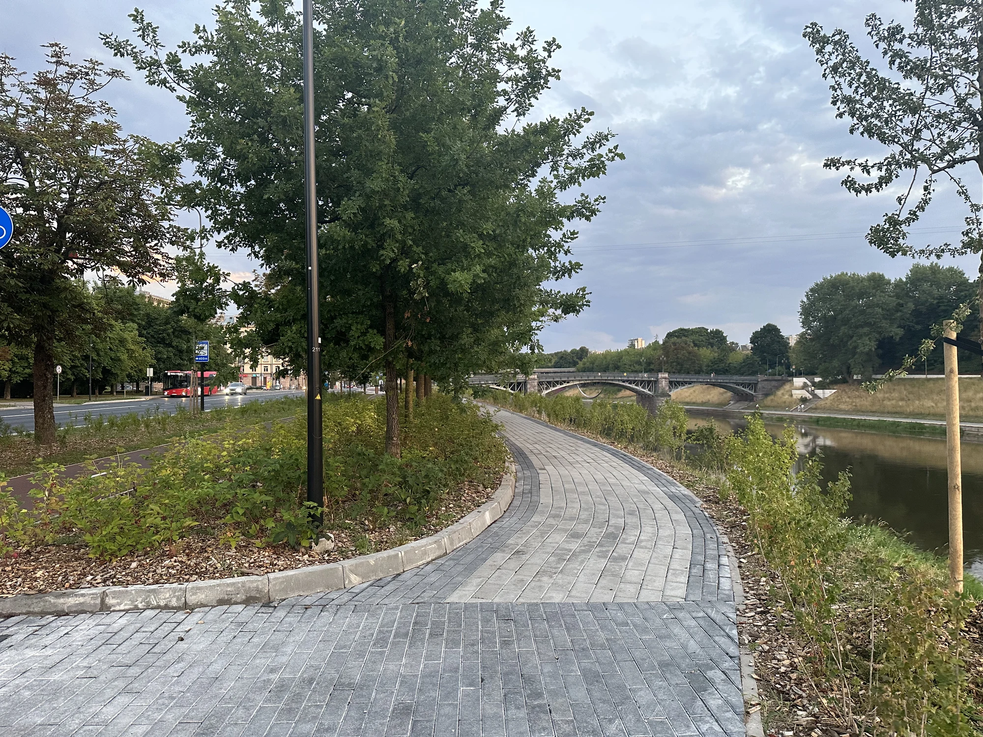A curved grey-paved pedestrian path along the Neris River in Vilnius, lined with green trees and leading towards a bridge under an overcast sky.