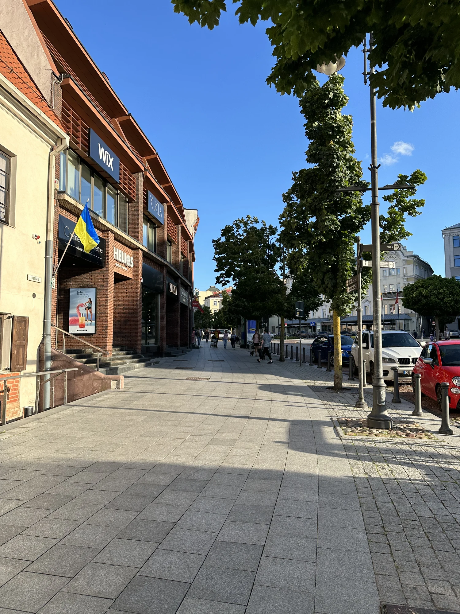 A wide paved sidewalk on Didžioji Street in Vilnius, lined with brick buildings, trees, parked cars, and pedestrians walking under a clear blue sky.