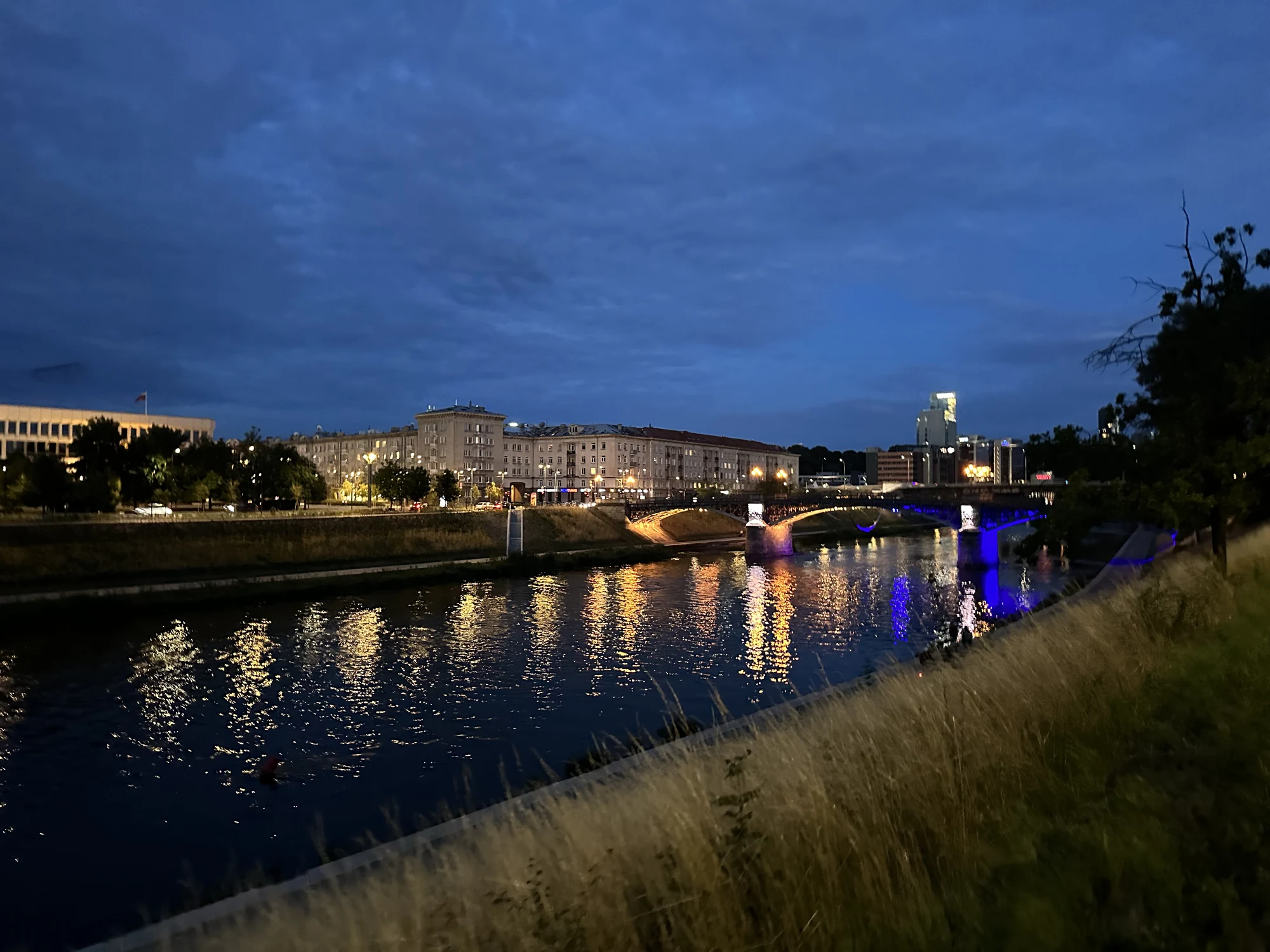 A twilight view of the Žvėrynas Bridge in Vilnius, illuminated with blue lights, reflecting on the Neris River with city buildings in the background.