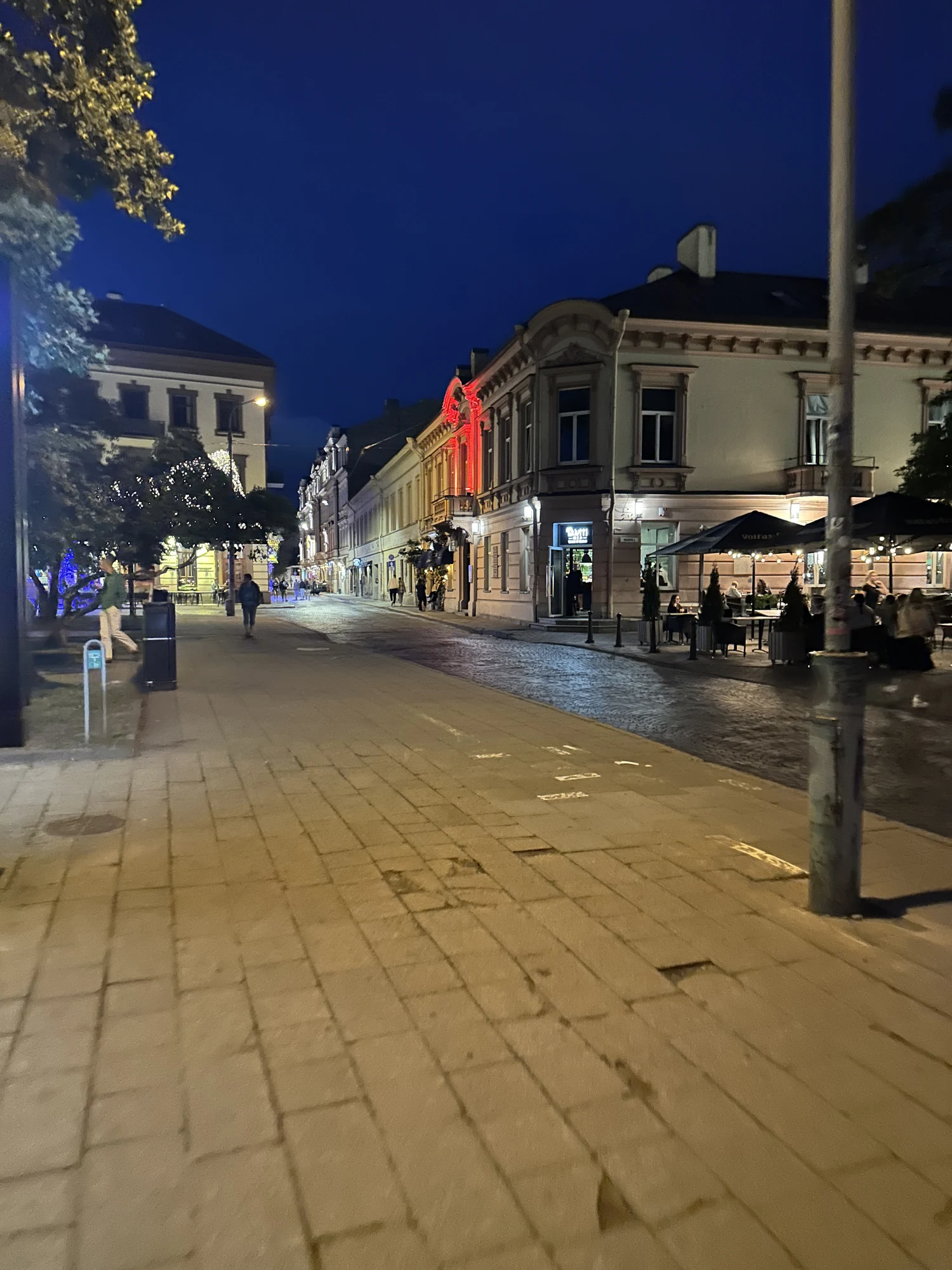 A pedestrian street in Vilnius Old Town at night, lined with historic buildings, outdoor cafe seating under umbrellas, and people walking.