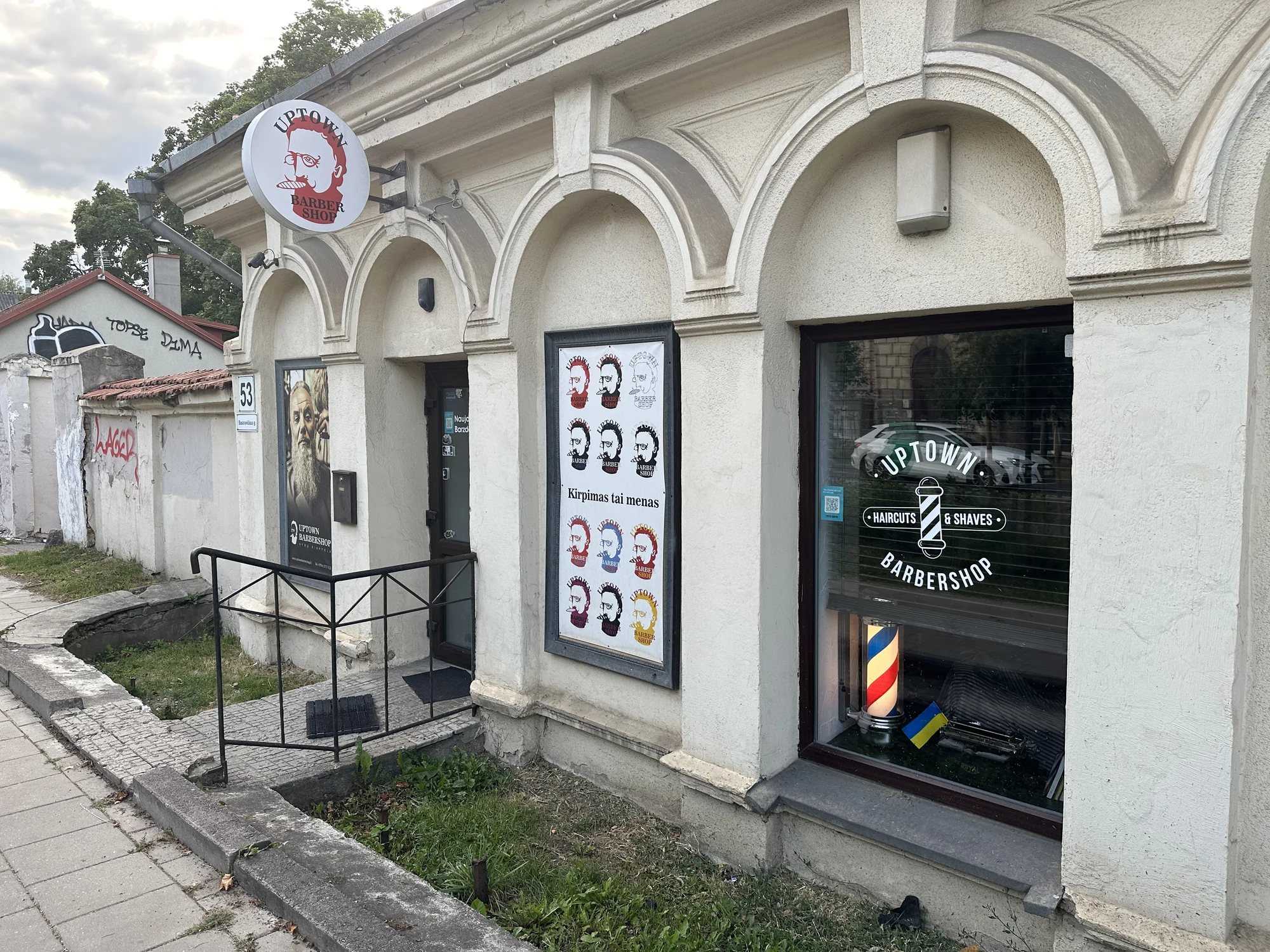 Exterior view of Uptown Barbershop in Vilnius, featuring arched windows, a circular logo sign, and posters on a light-colored stone building.