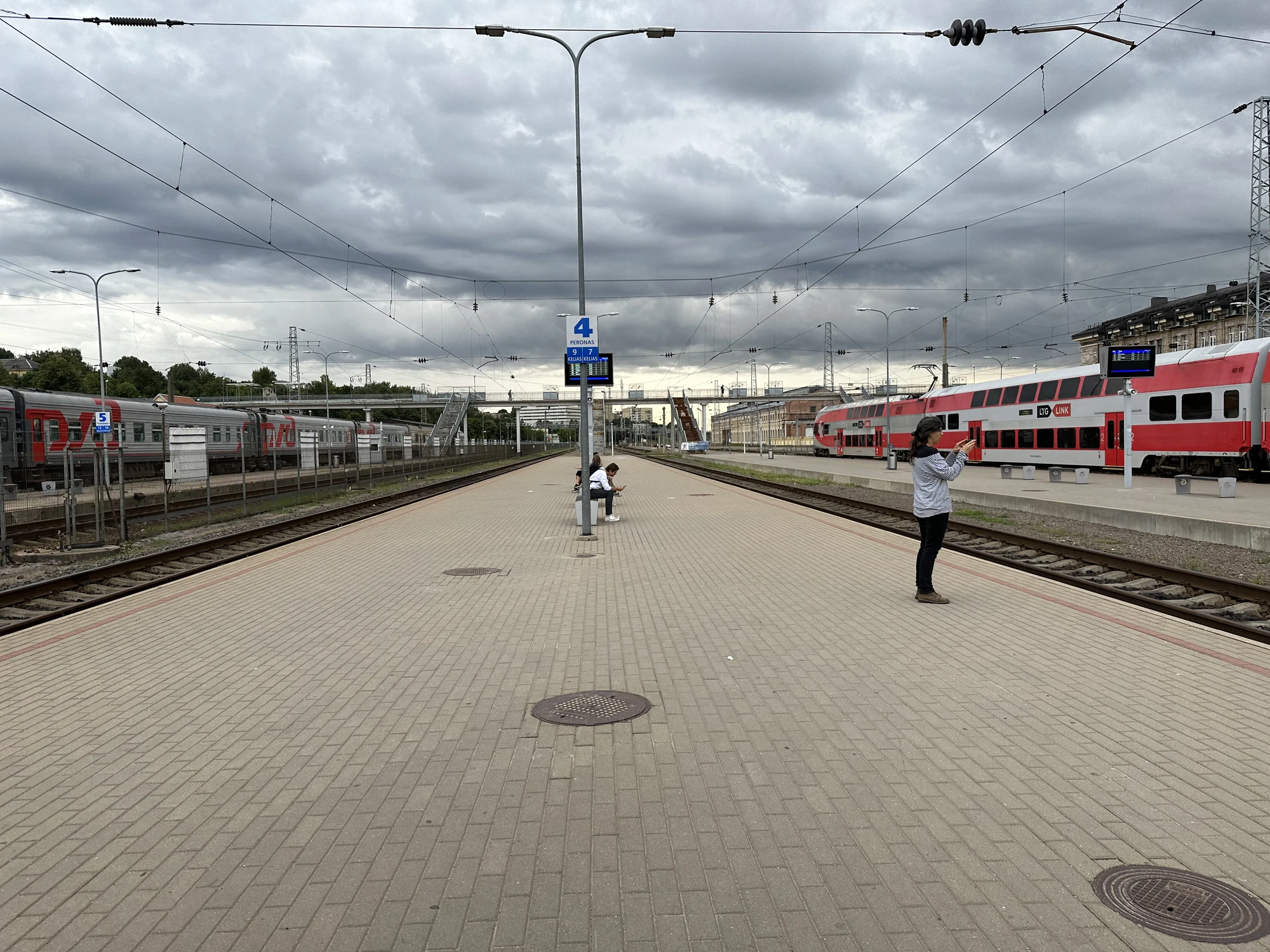 A wide paved railway platform at Vilnius Railway Station with a double-decker red train on the right and a grey train on the left under an overcast sky.