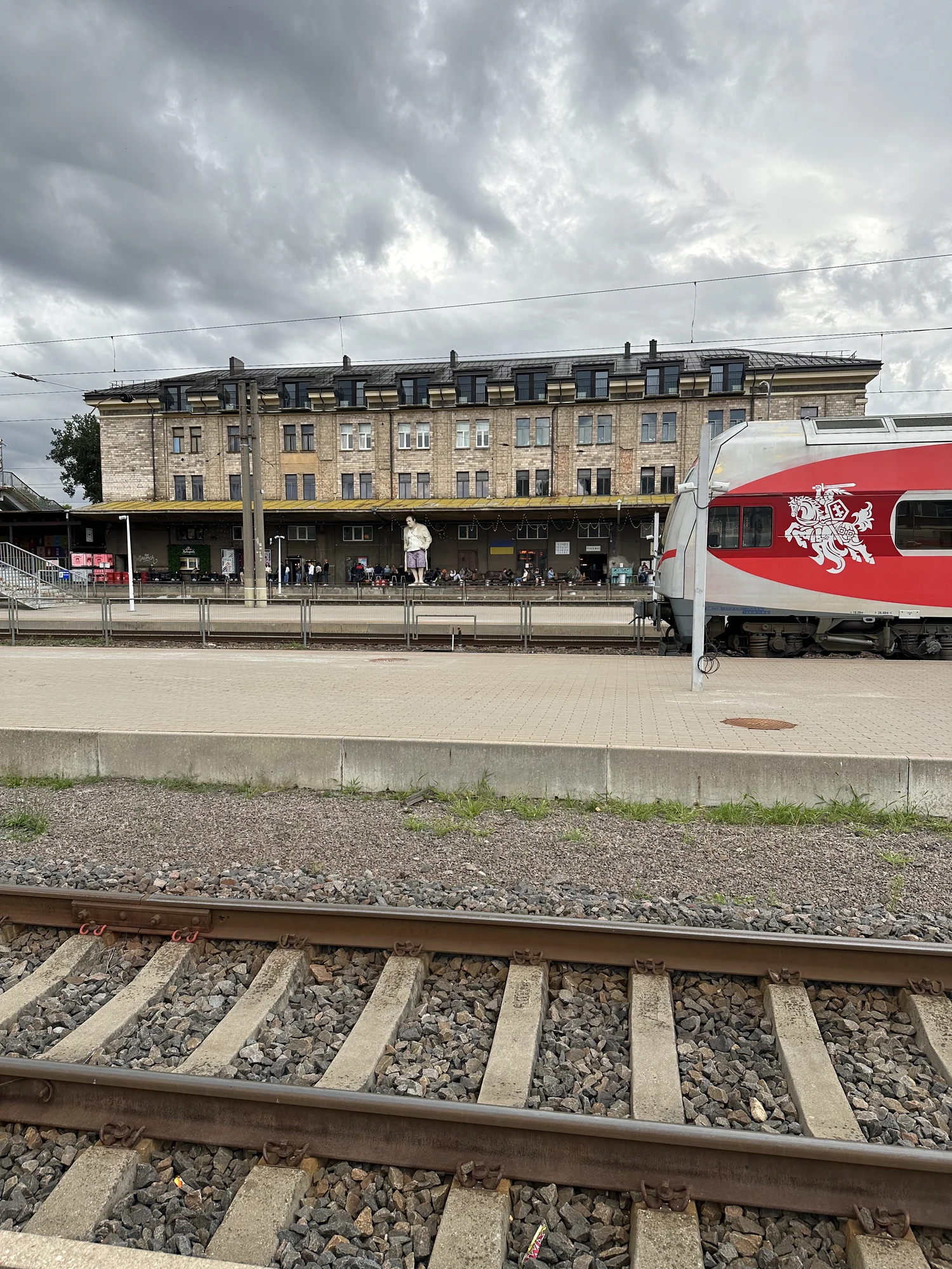 A view across train tracks at the Vilnius Railway Station, showing a red and silver Lithuanian train on the right and a large statue of Tony Soprano on the platform in front of a historic stone building.