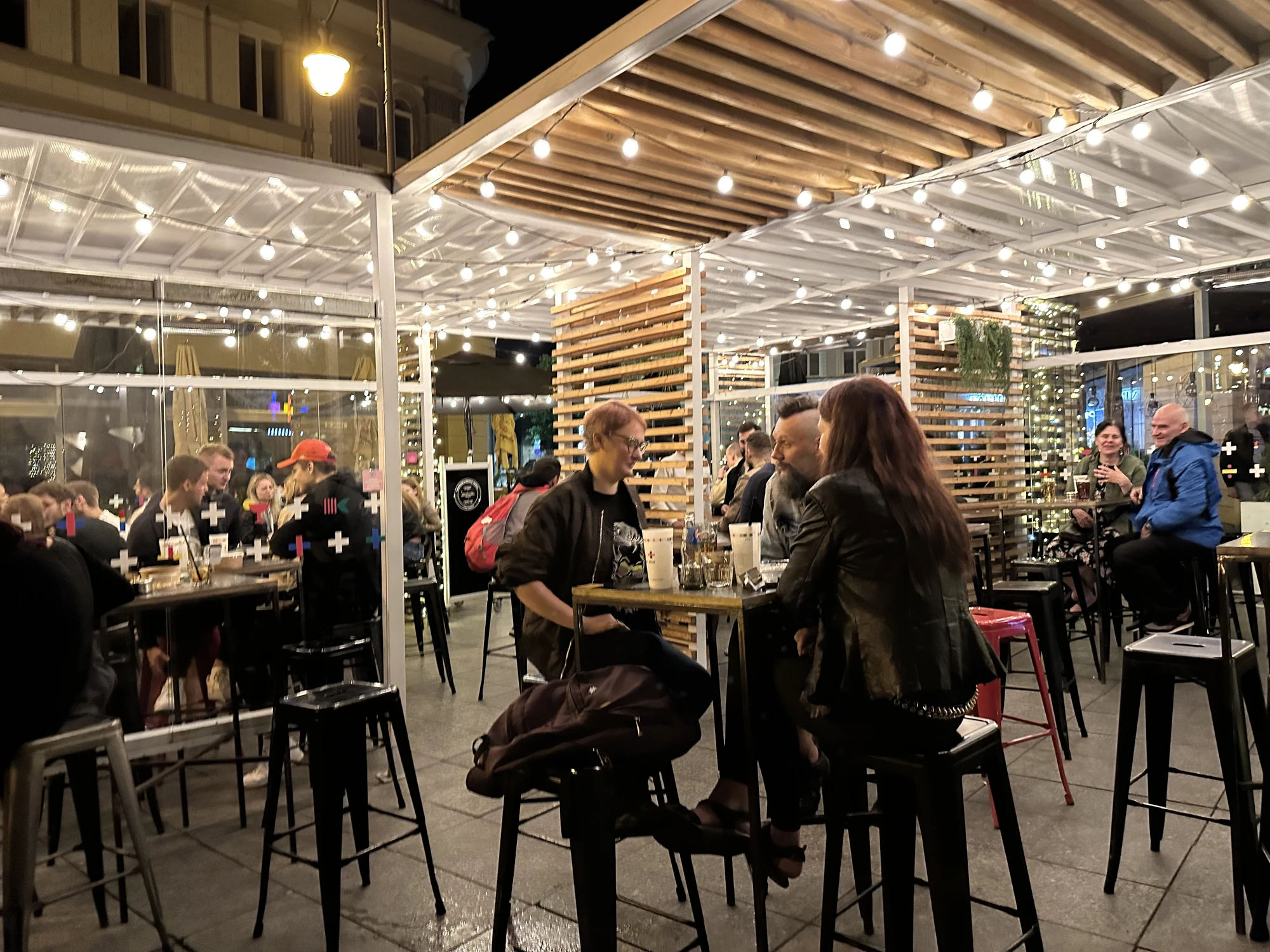 A lively outdoor patio bar at night in Vilnius, featuring people sitting at high tables under a wooden pergola decorated with warm string lights.