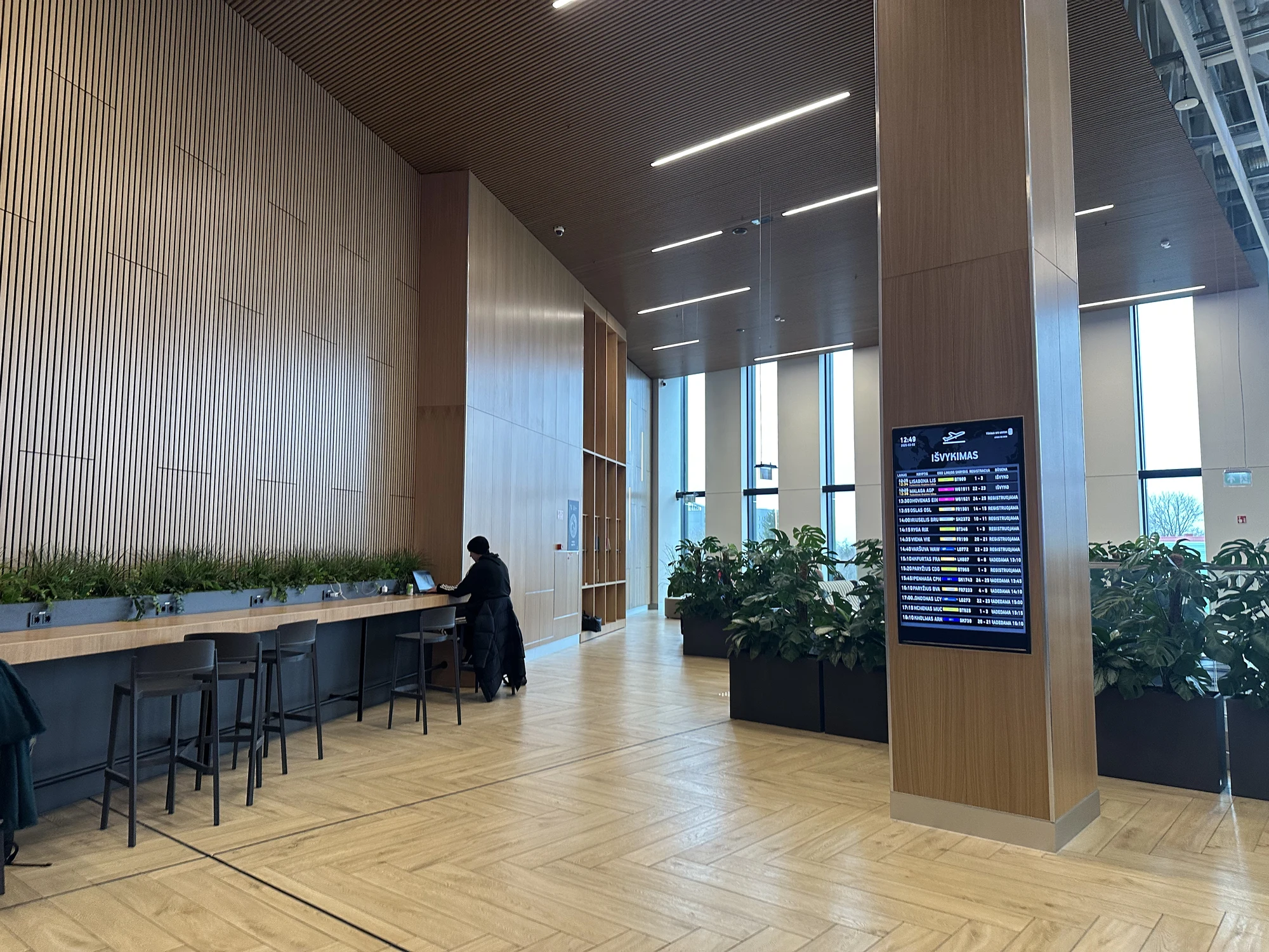 A modern airport terminal interior with light wood paneled walls and pillars, a long work counter with bar stools, green potted plants, and a digital departures board in Lithuanian.