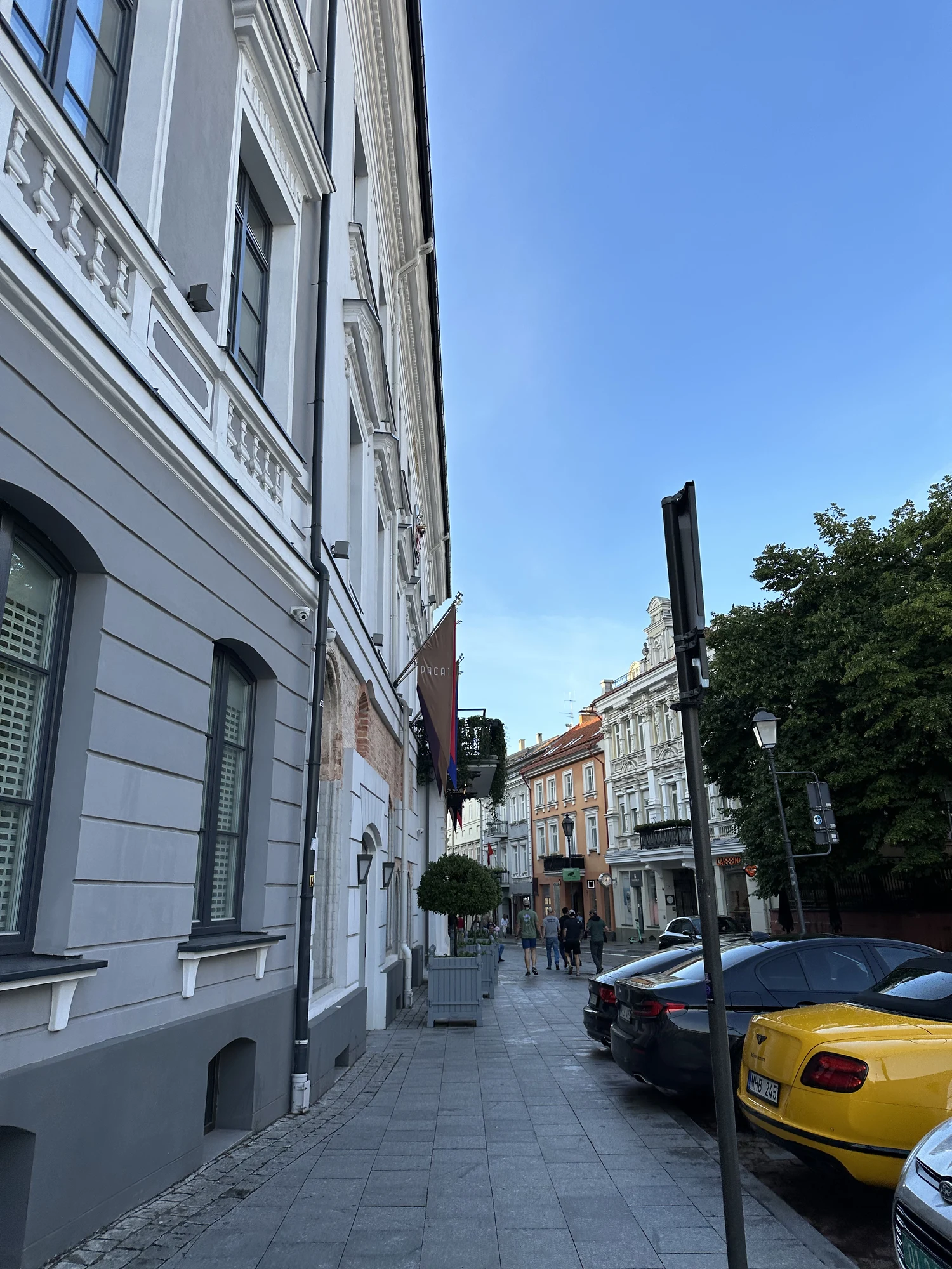 A view looking down a paved sidewalk on Didžioji Street in Vilnius, featuring the grey facade of Hotel Pacai on the left and parked cars on the right under a clear blue sky.