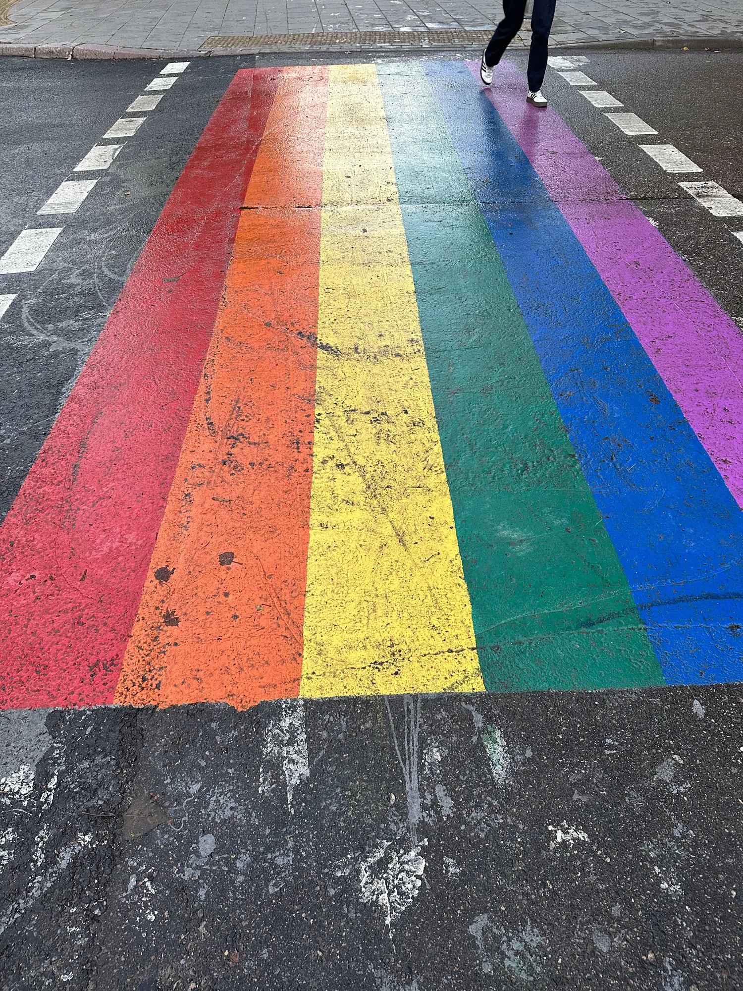 A high-angle shot of a pedestrian crosswalk painted in the bright colors of the rainbow pride flag, with a person's legs visible walking across the top right.