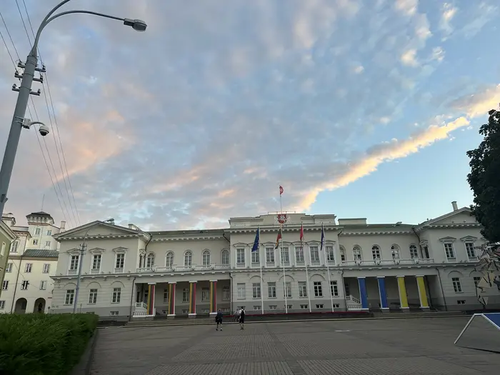 The neoclassical facade of the Presidential Palace of Lithuania in Vilnius, viewed from Daukanto Square under a sky with pink-tinged clouds.