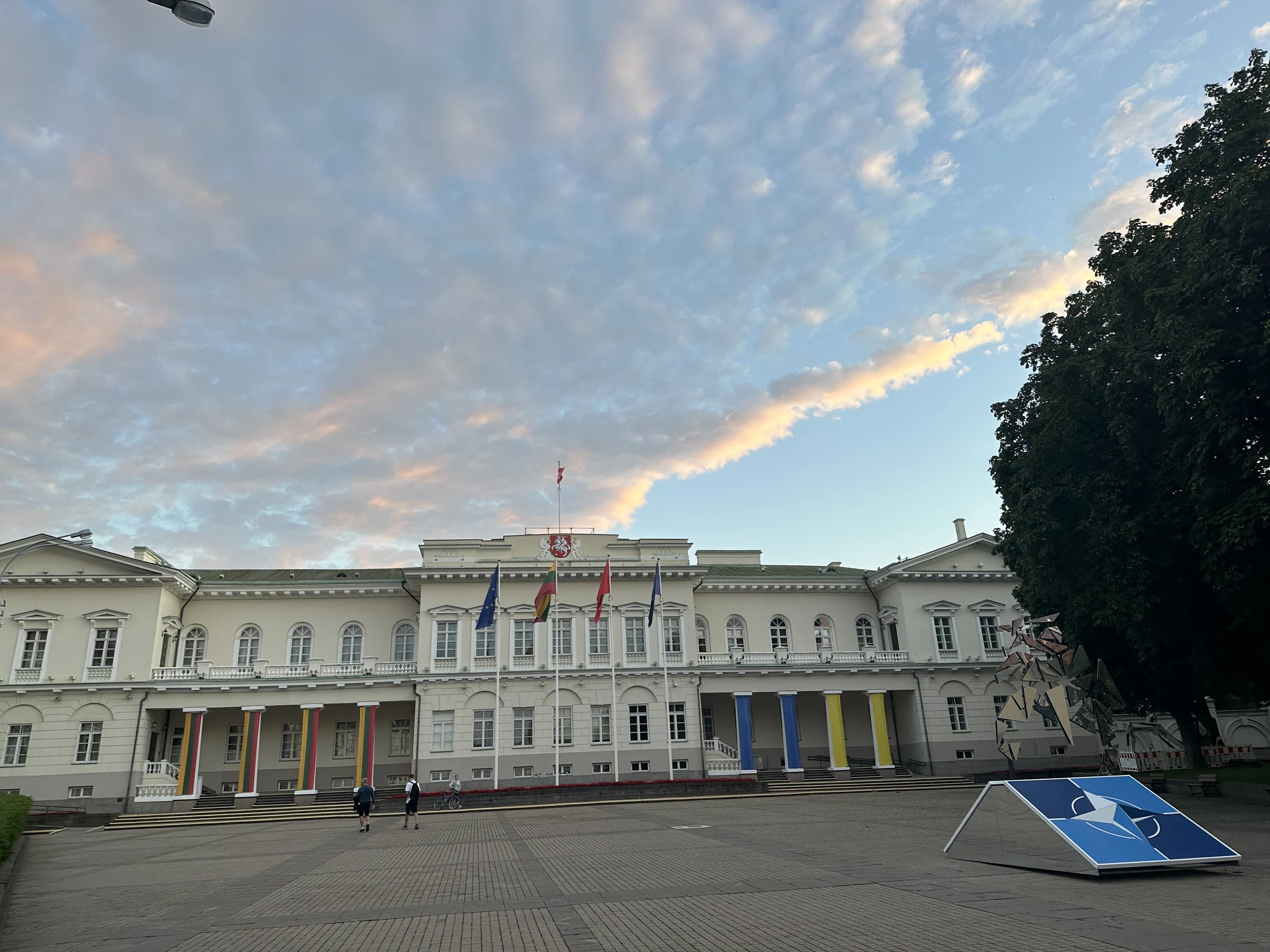 The white neoclassical facade of the Presidential Palace of Lithuania in Vilnius, seen from Daukanto Square under a cloudy sunset sky.