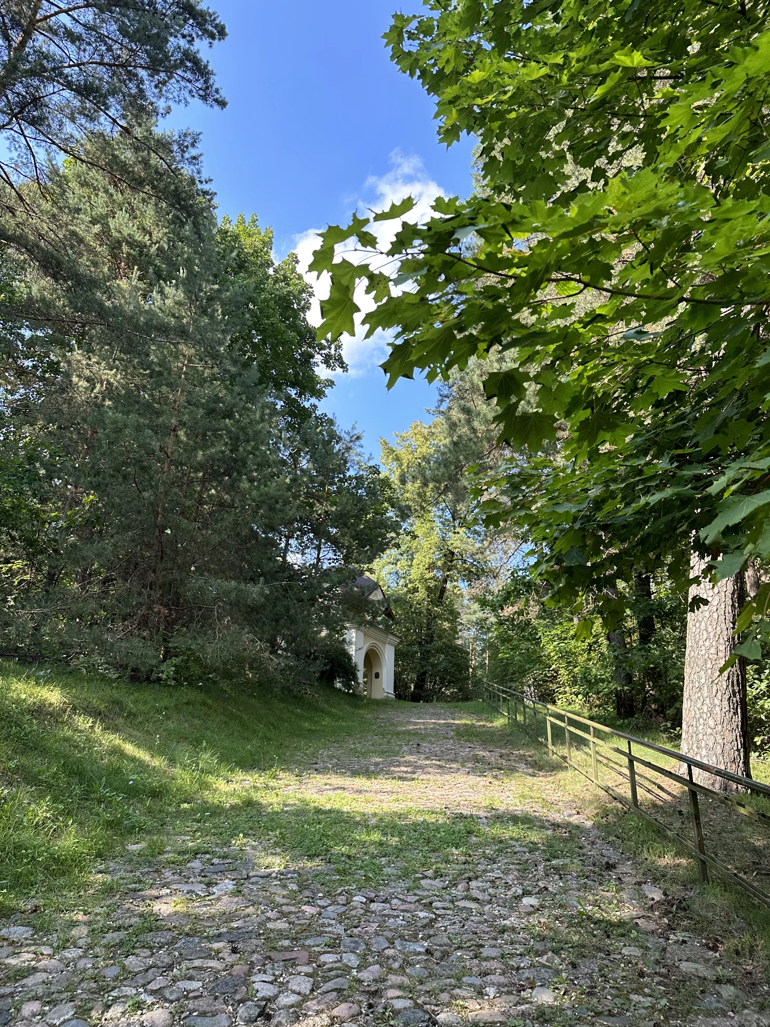 A stone-paved path climbs a hill flanked by green trees and a metal handrail, leading towards a small white chapel under a blue sky.