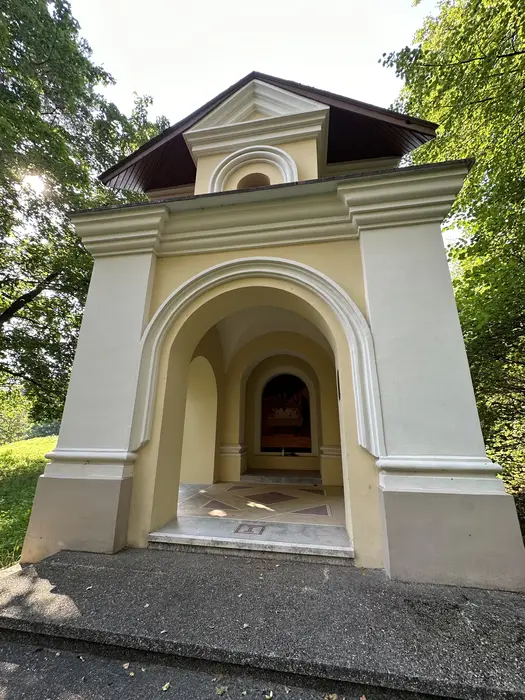 A yellow and white baroque-style chapel with arched openings, part of the Vilnius Calvary Stations of the Cross, surrounded by green trees.