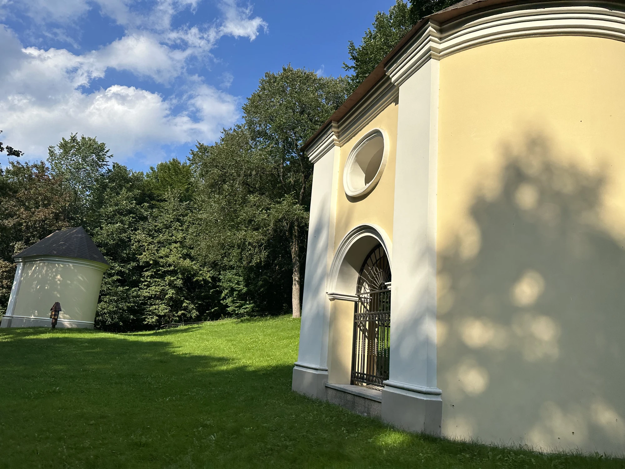 A yellow and white baroque chapel on a grassy hill with another smaller chapel in the background surrounded by trees.