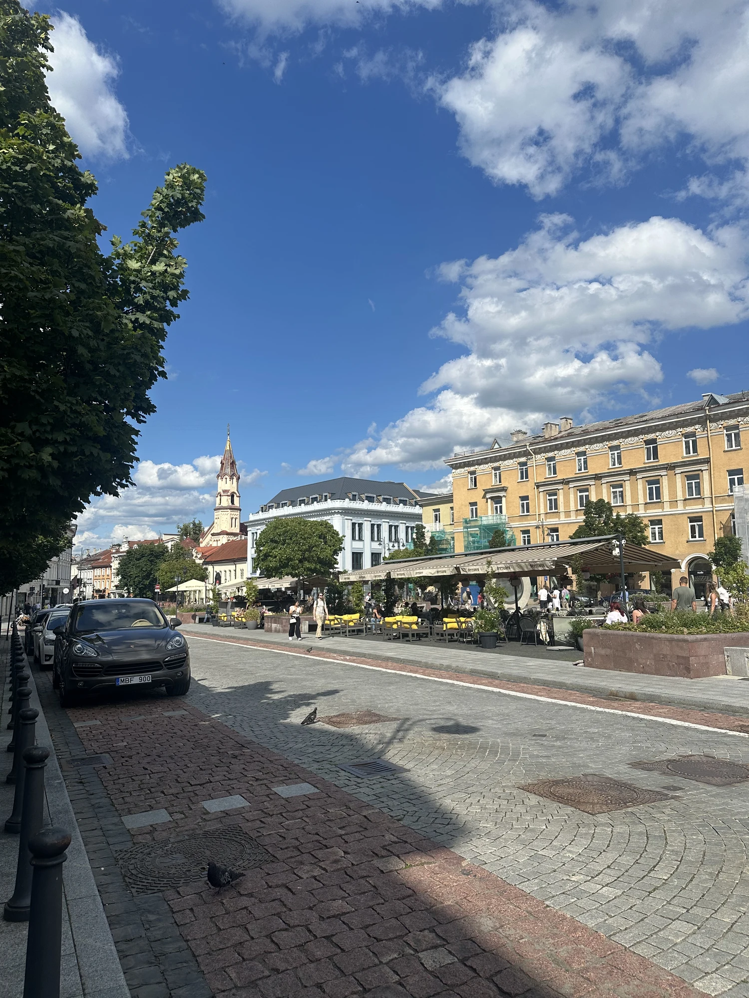A cobblestone street in Vilnius Old Town with outdoor cafe seating, historic buildings, a church spire in the distance, and a parked car on the left under a blue sky with white clouds.