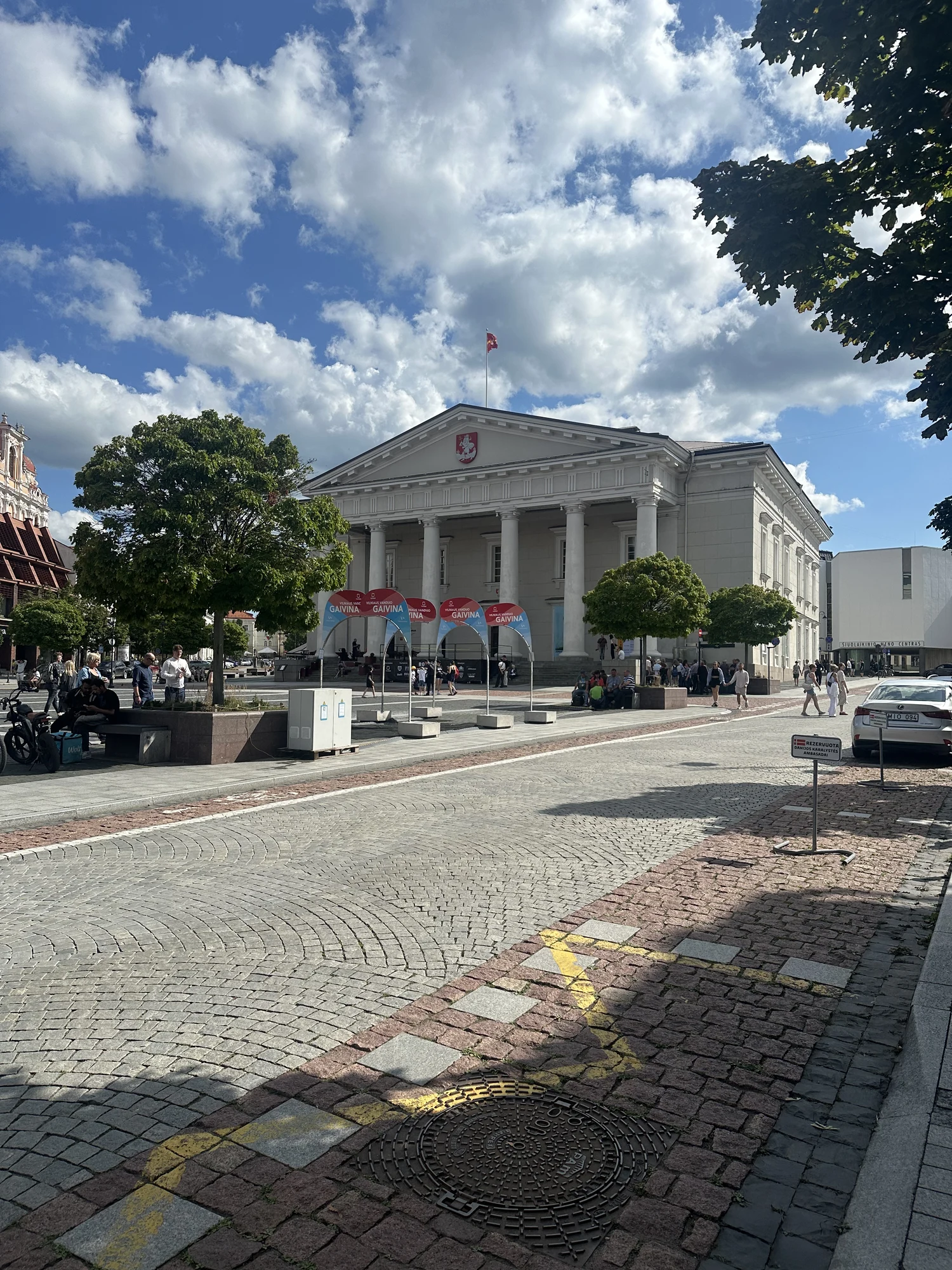 A sunny outdoor shot of the neoclassical Vilnius Town Hall building with its grand columns, facing a cobblestone square with pedestrians and green trees.