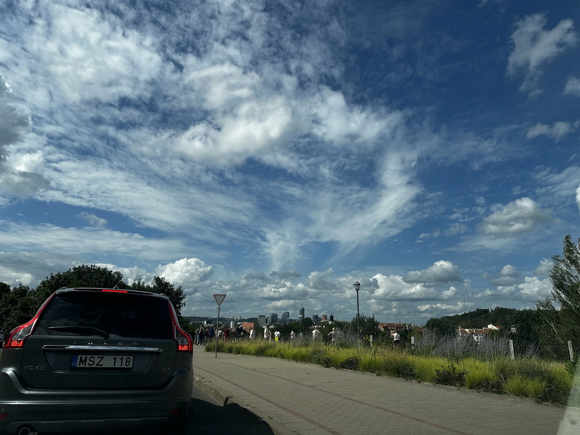 A view from a car window showing a grey Volvo parked on the left, a paved path, and a distant view of the Vilnius modern skyline under a bright blue sky filled with wispy white clouds.