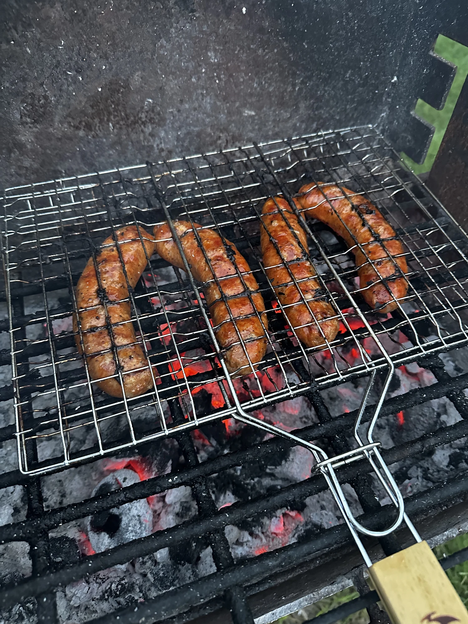 Four sausages inside a metal grilling basket cook over glowing red charcoal embers on an outdoor grill.