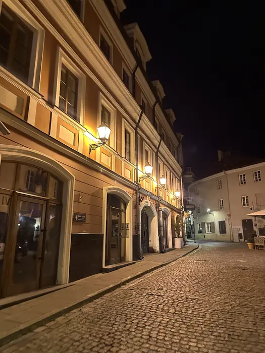 A quiet cobblestone street in Vilnius Old Town at night, lined with historic buildings illuminated by warm wall-mounted street lamps.