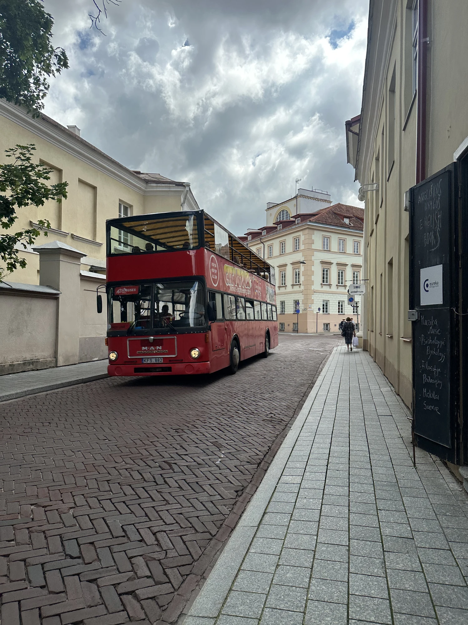 A red double-decker tour bus driving down a narrow cobblestone street lined with historic yellow buildings under a cloudy sky in Vilnius.