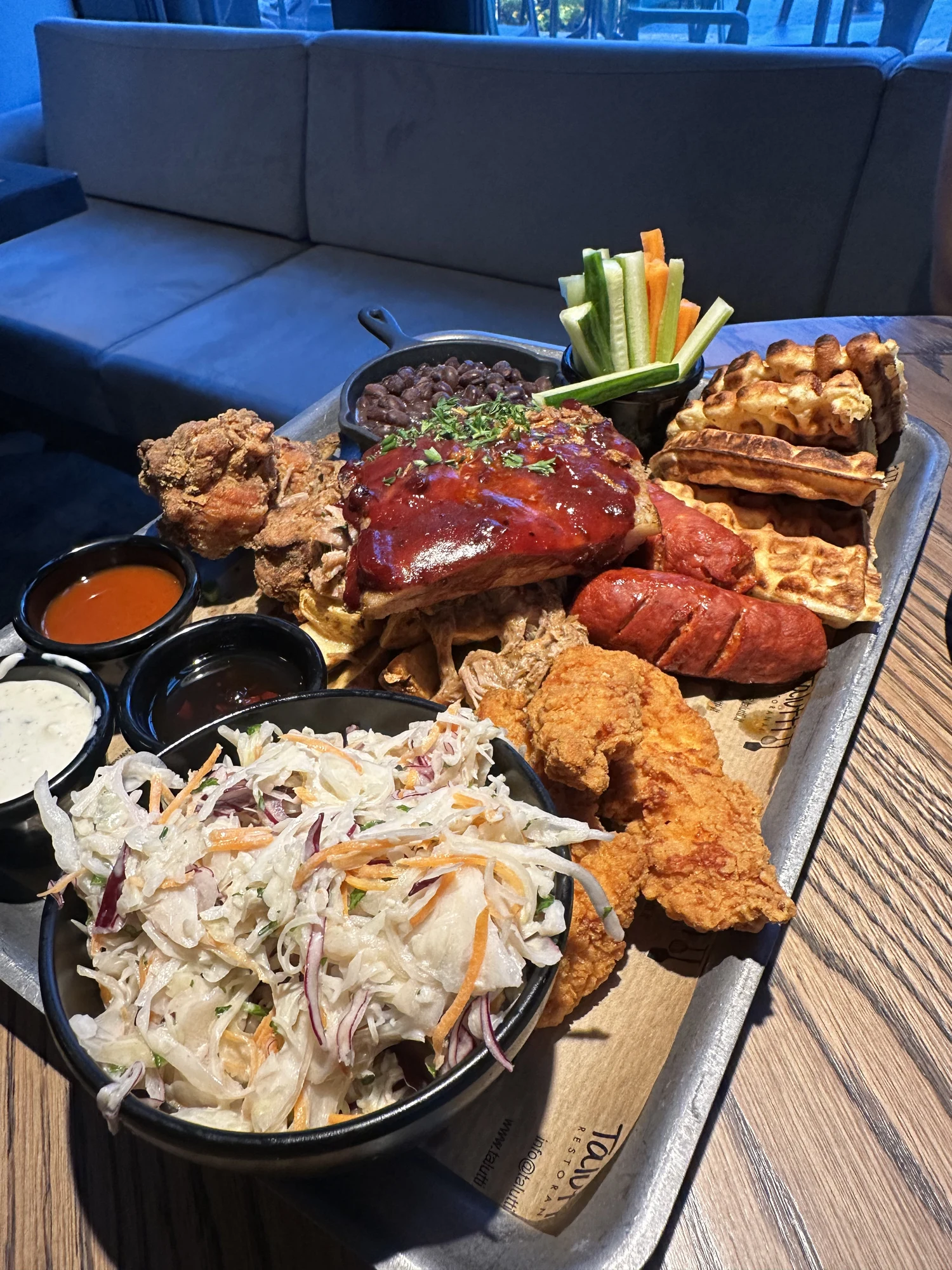 A large metal tray filled with BBQ ribs, fried chicken tenders, waffles, sausages, coleslaw, black beans, and dipping sauces on a wooden table.