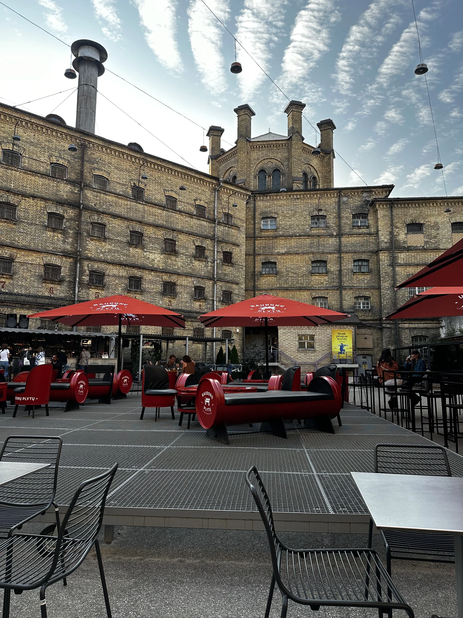 An outdoor patio with red umbrellas and barrel-shaped benches in the courtyard of the historic yellow-brick Lukiškės Prison in Vilnius.