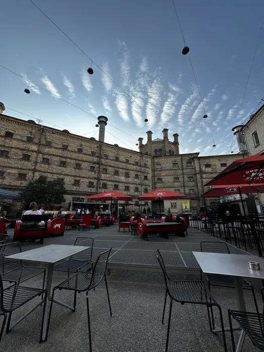 An outdoor cafe area with black chairs, tables, and red umbrellas set up in the courtyard of the historic brick Lukiškės Prison building under a blue sky with wispy clouds.