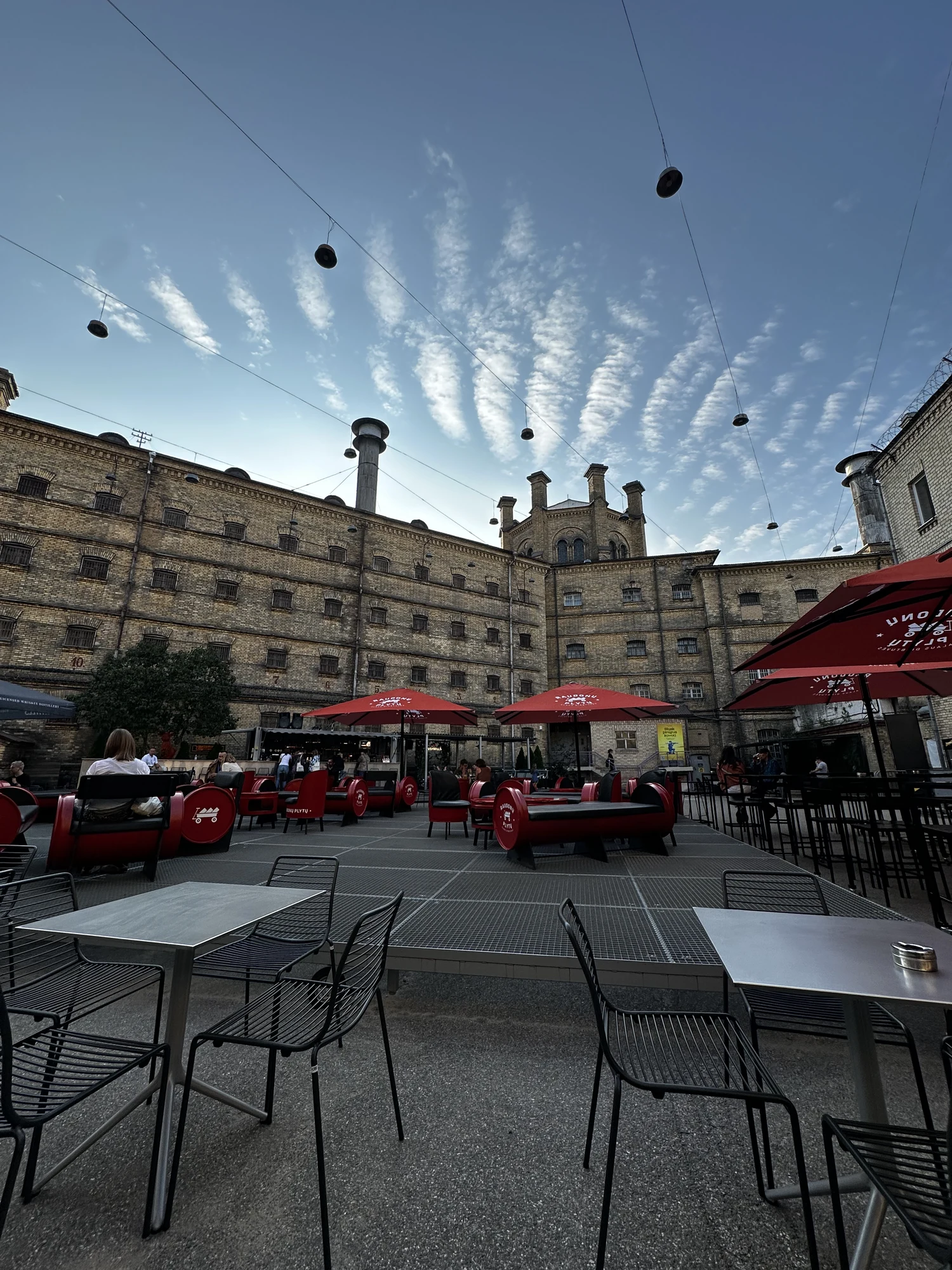 An outdoor cafe area with black chairs, tables, and red umbrellas set up in the courtyard of the historic brick Lukiškės Prison building under a blue sky with wispy clouds.