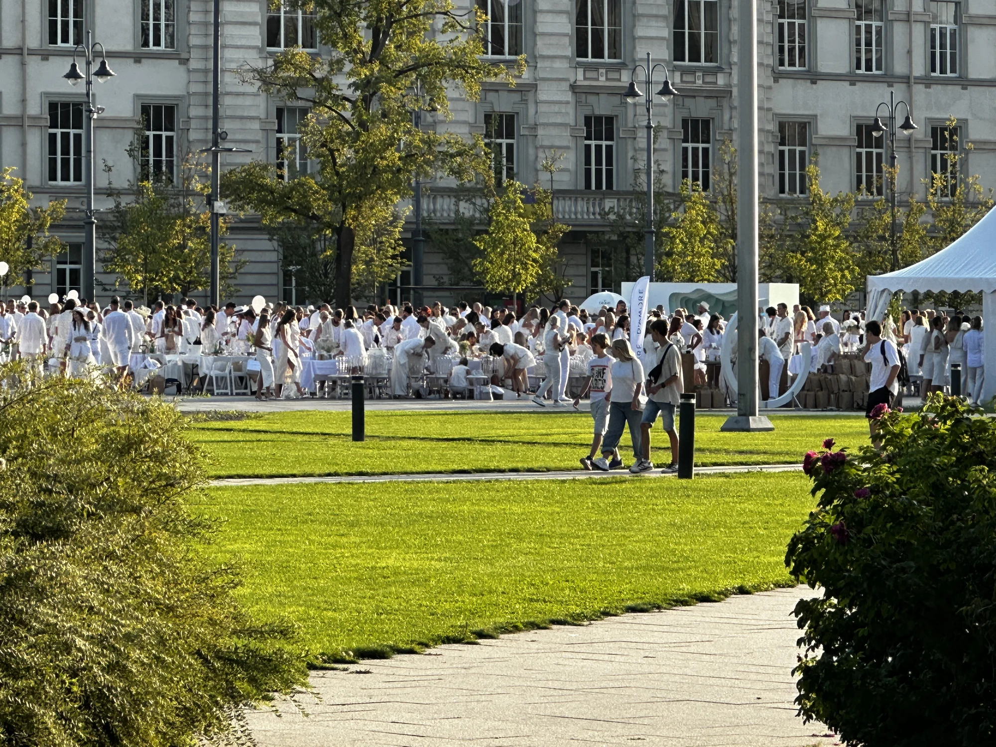 A large crowd of people dressed entirely in white gathered for an outdoor dinner event on a green lawn in front of a grand historic building in Vilnius.