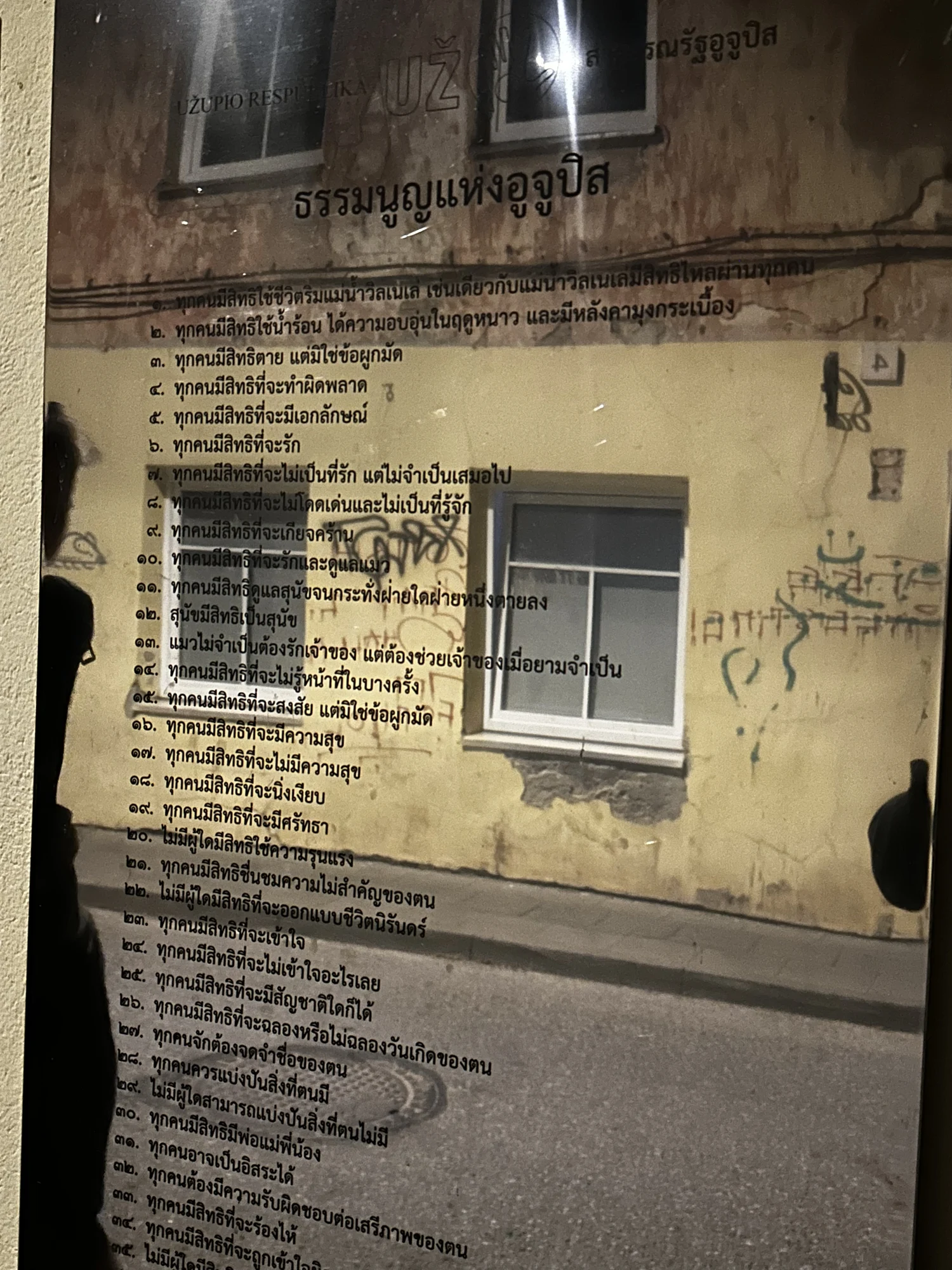 A reflective metal plaque on an outdoor wall displaying the Constitution of the Republic of Užupis written in Thai script.