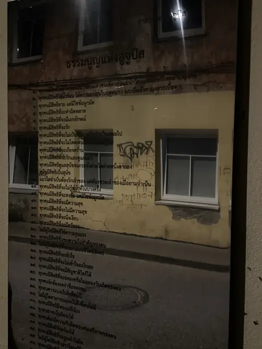 A reflective plaque on an outdoor wall displaying the Constitution of Užupis written in Thai script, reflecting the street and building opposite.