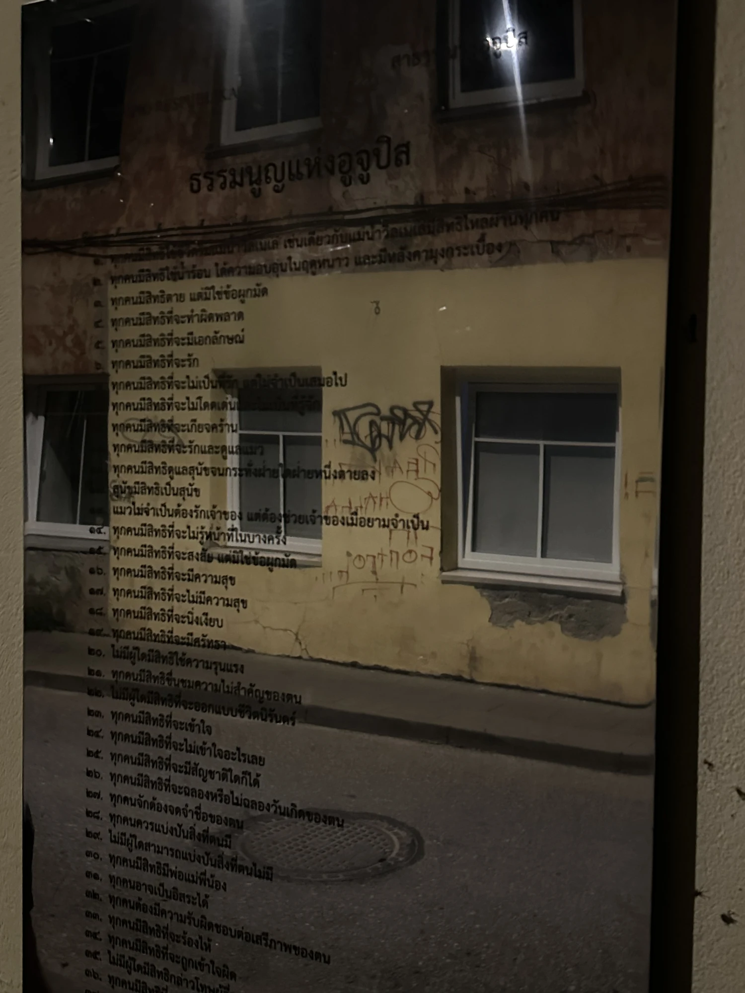 A reflective plaque on an outdoor wall displaying the Constitution of Užupis written in Thai script, reflecting the street and building opposite.
